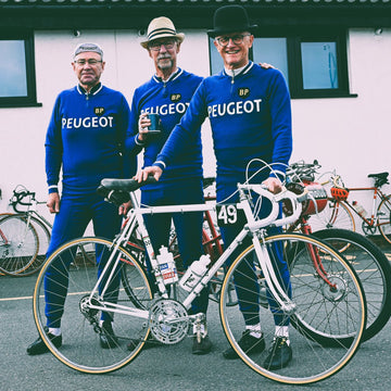 A group of smartly dressed men in wearing retro blue Peugeot team tracksuits with a Peugeot team bike at the Simpson Retro 2024.