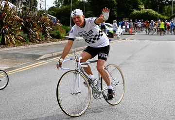 A man in retro Peugeot team kit riding a Peugeot bike with a grey frame - he's waving to the camera.