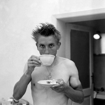 Candid picture of Jacques Anquetil drinking a coffee, with wild hair as though he has just got out of the shower. 